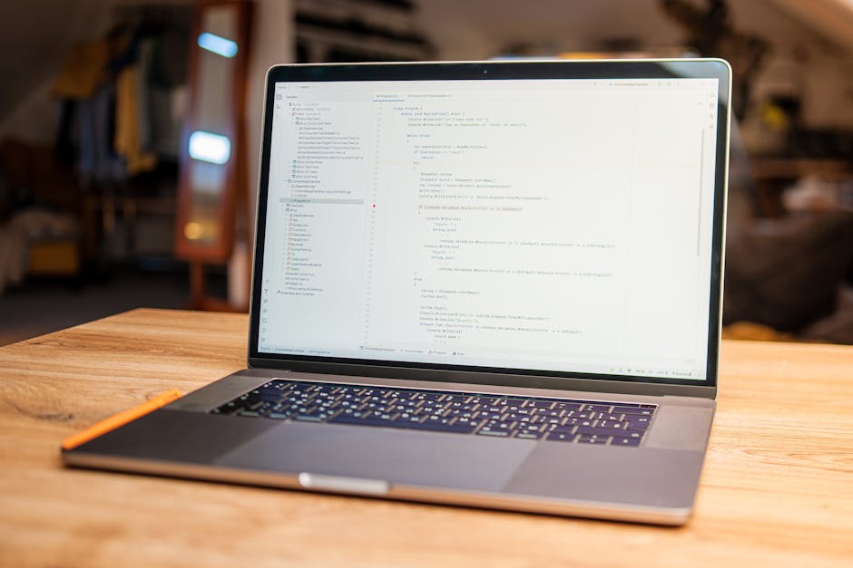 A person typing on a laptop with a black keyboard and a slim profile, positioned on a flat white surface, displaying a coding or programming interface on the screen. The screen shows multiple lines of
