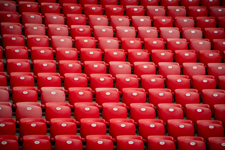 A large array of bright red plastic stadium seats arranged in multiple horizontal rows, each seat featuring a small white sticker with black alphanumeric codes such as 'DN-29', 'DR-36', and 'DQ-35'. T
